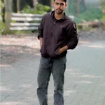 A young man stands casually outdoors on a paved path, wearing a dark jacket, jeans, and sneakers, embodying confidence and style—the freelance digital marketing analyst in Kannur.