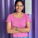A confident woman stands with folded arms against a vibrant purple wall, dressed in a pink top and blue jeans, representing the best freelance digital marketing strategist in Kannur.
