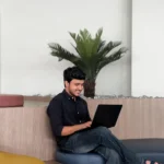 A man in a dark shirt and jeans sits comfortably with a laptop in a modern space featuring a small palm plant, reflecting the best freelance digital marketing analyst in Kannur.