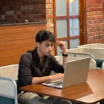 A man in a black shirt works intently on a laptop at a cozy cafe with brick walls, embodying the freelance digital marketing analyst in Kannur.
