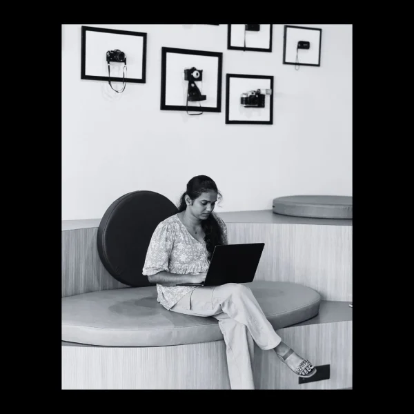 A freelance digital marketing analyst in Kannur sits on a modern circular bench, working on a laptop. The minimalist room features framed vintage cameras displayed on a white wall in the background. The photo is in black and white.